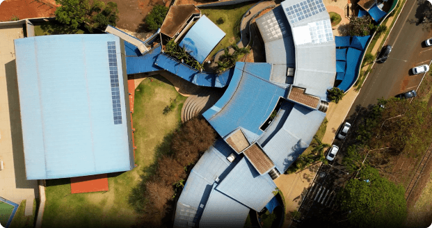 Vista aérea do Educativa Centro Educacional, mostrando a arquitetura moderna e curvilínea dos prédios com telhados azuis e painéis solares, rodeados por áreas verdes e ruas arborizadas.